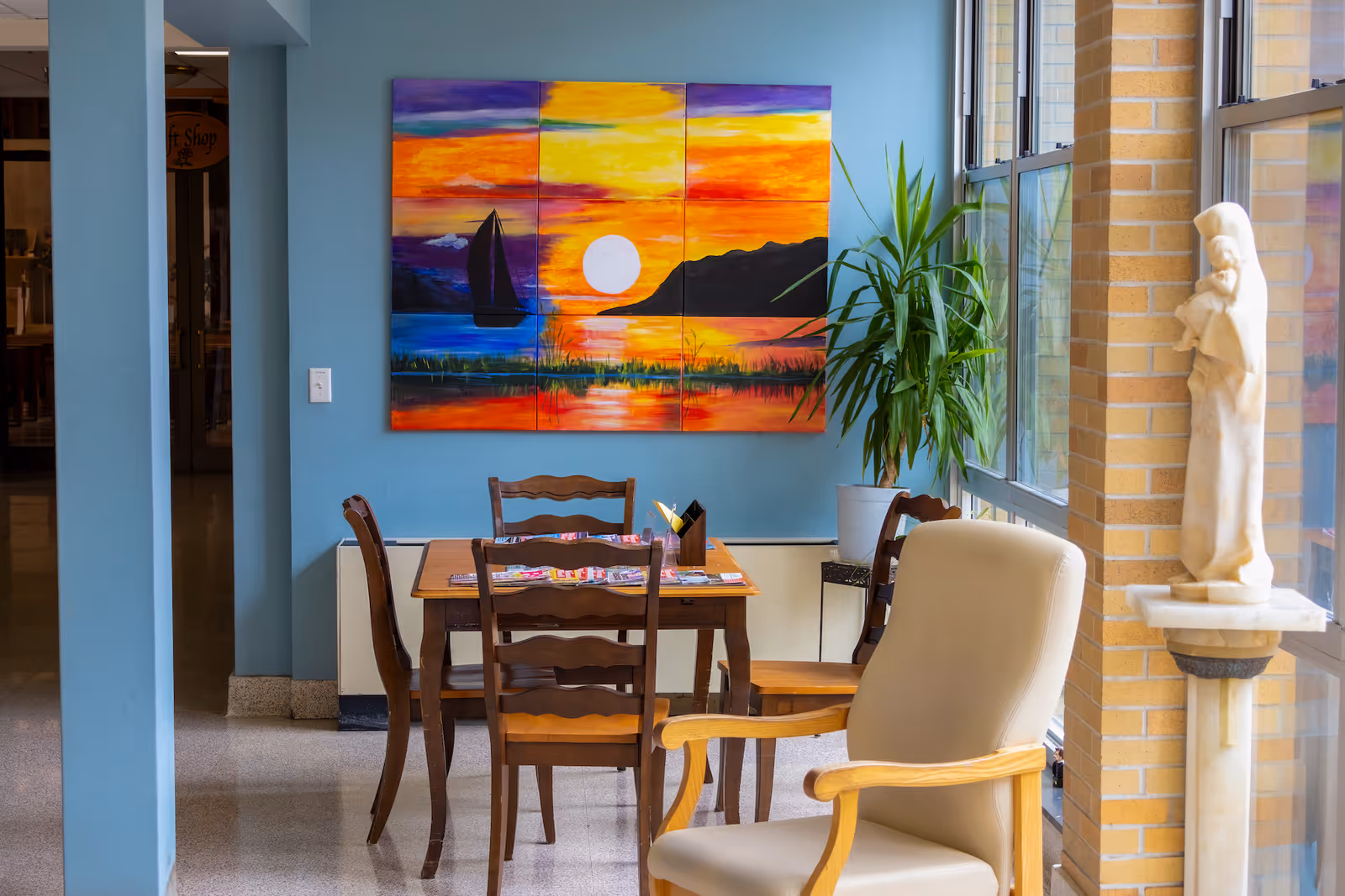 A cozy seating area in a senior living facility with a wooden table and four chairs, a beige armchair, a large colorful painting of a sunset with a sailboat on the wall, a potted plant, and a statue near large windows letting in natural light.