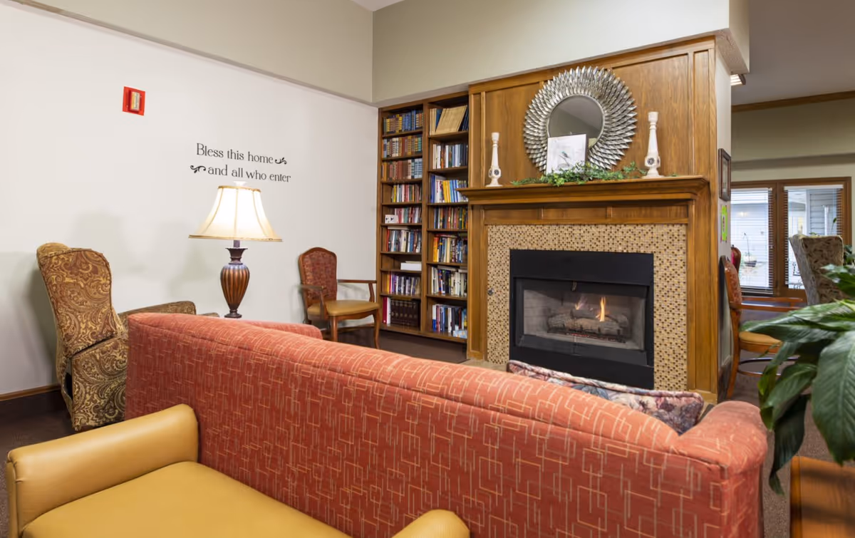 A cozy living room area with a red patterned sofa and a yellow armchair facing a fireplace with a decorative mirror and candlesticks on the mantel. To the left, there is a tall bookshelf filled with books and a lamp on a side table between two chairs. A wall decal reads 'Bless this home and all who enter.'