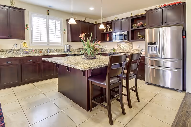 Bright modern kitchen with a central island, granite countertops, dark wood cabinets, a stainless steel refrigerator, and two bar stools.