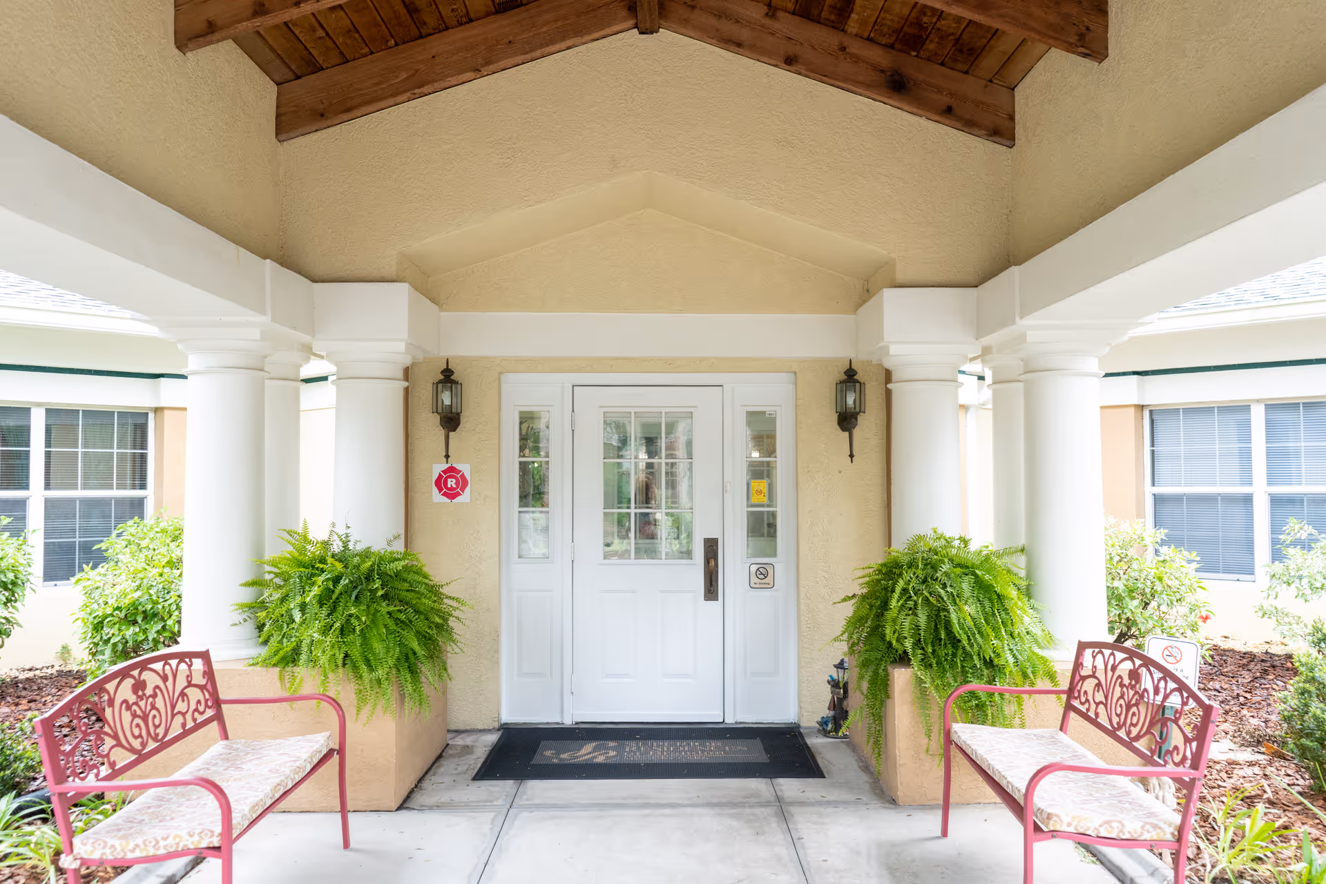 Covered entrance to a senior living facility with white double doors, columns, potted ferns, and benches.