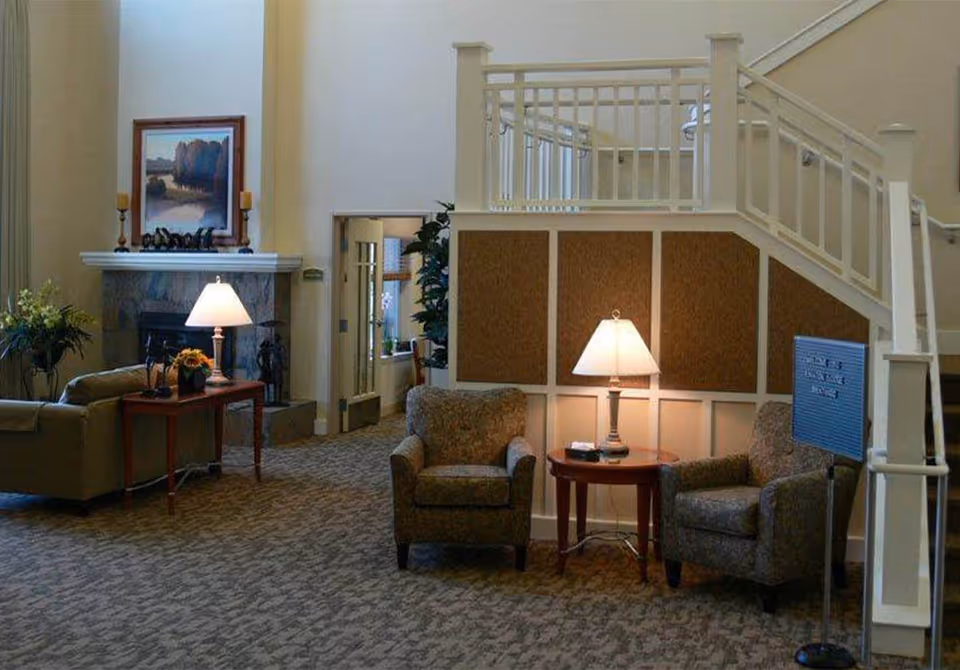 A senior living lounge with armchairs and side tables illuminated by lamps, a fireplace, plants, and an interior staircase railing.