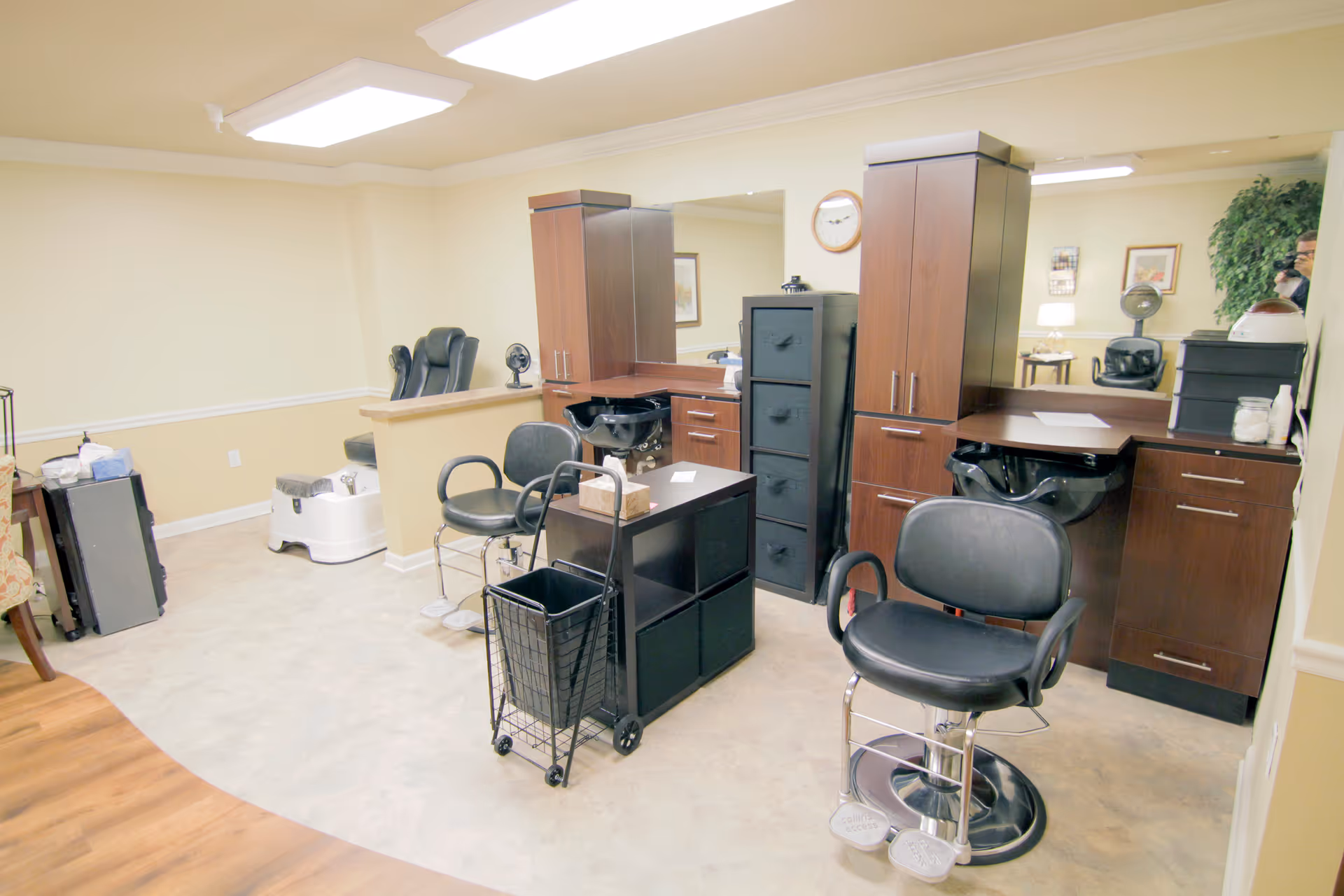 Interior view of a salon area in a senior living facility with two black salon chairs, a pedicure station, wooden cabinets with a large mirror, and various salon supplies. The room has light-colored walls and flooring with a wooden floor section visible.