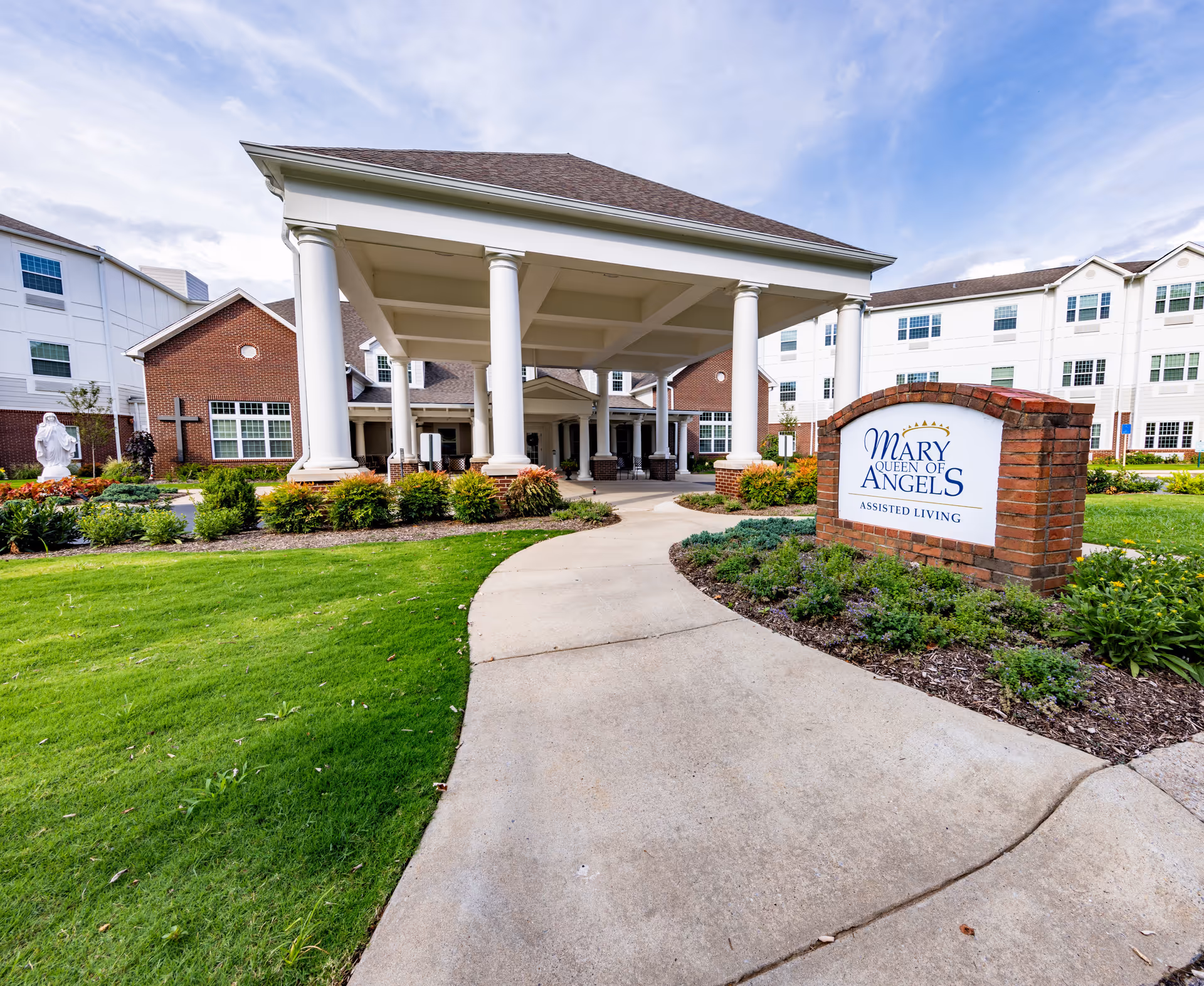 Entrance of Mary Queen of Angels Assisted Living facility with a covered driveway supported by white columns, a brick sign displaying the facility name, well-maintained green lawn, landscaped bushes, and a statue in the garden area.