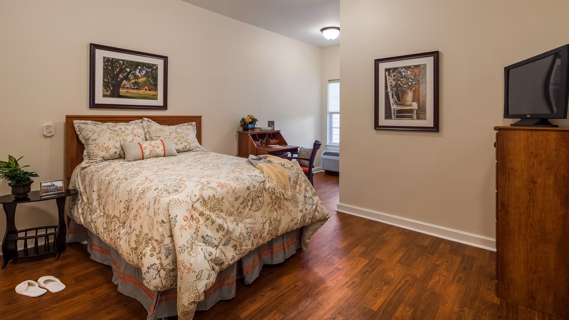 A cozy bedroom with a wooden bed frame and floral patterned bedding. There is a small side table with a plant and a framed photo, a wooden desk with a chair, and a wooden dresser with a TV on top. The room has hardwood floors, two framed pictures on the walls, and a window letting in natural light.