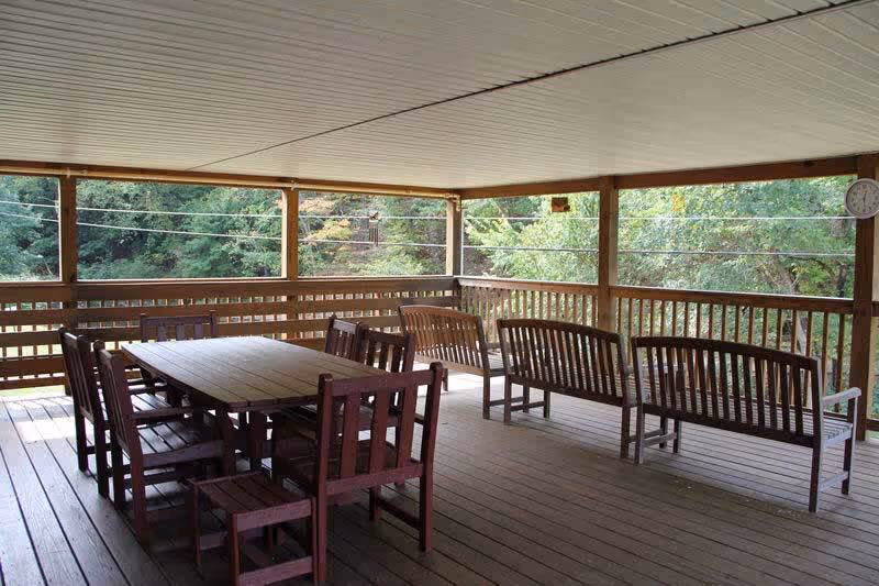 Covered outdoor wooden deck with a rectangular wooden table surrounded by eight wooden chairs on the left side and three wooden benches on the right side. The deck is surrounded by wooden railings and overlooks green trees and foliage.