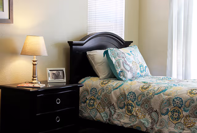 A neatly made single bed with patterned bedding and pillows beside a dark wood nightstand holding a lamp and framed photo in a bright bedroom.