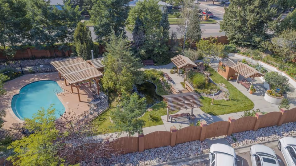 Aerial view of an outdoor garden area at Medallion Post Acute Rehabilitation featuring a small swimming pool, several wooden pergolas with benches, a pond surrounded by greenery, and a walking path. The area is enclosed by a wooden fence and bordered by parked cars.