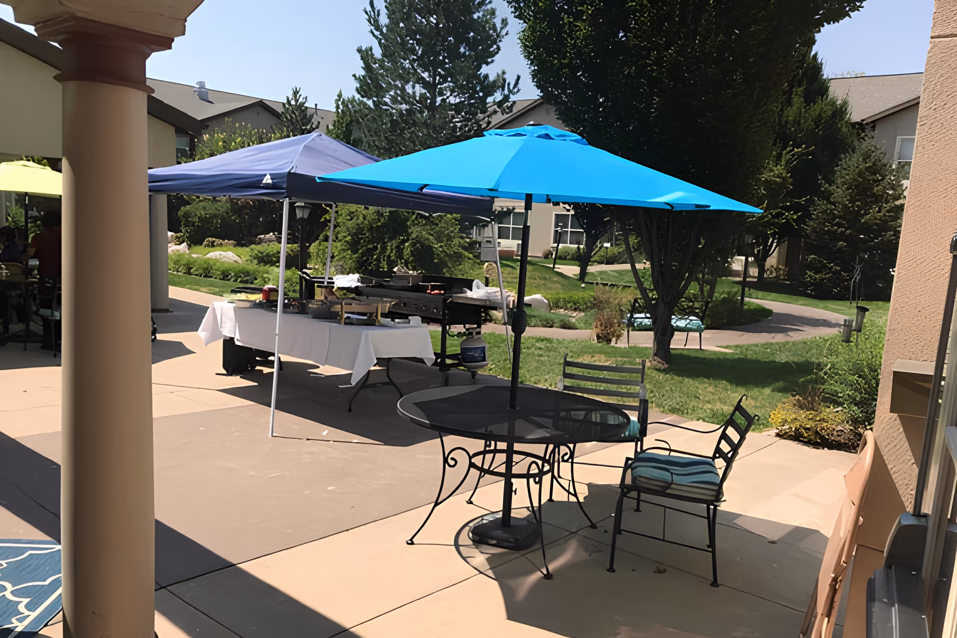 Outdoor patio area at Avamere at Mountain Ridge with a round metal table and two chairs under a bright blue umbrella. Nearby, there is a canopy tent with a table covered in a white tablecloth and a grill behind it. The background shows green grass, trees, and parts of the facility buildings under a clear sky.