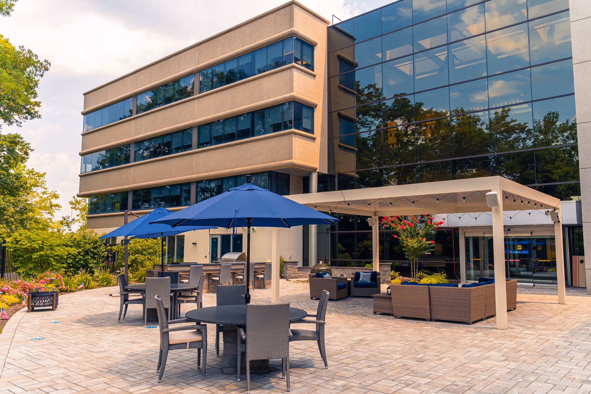 Outdoor patio area of a senior living facility with several round tables and chairs under blue umbrellas, a covered seating area with wicker furniture and cushions, surrounded by greenery and flowers, adjacent to a modern building with large reflective windows.