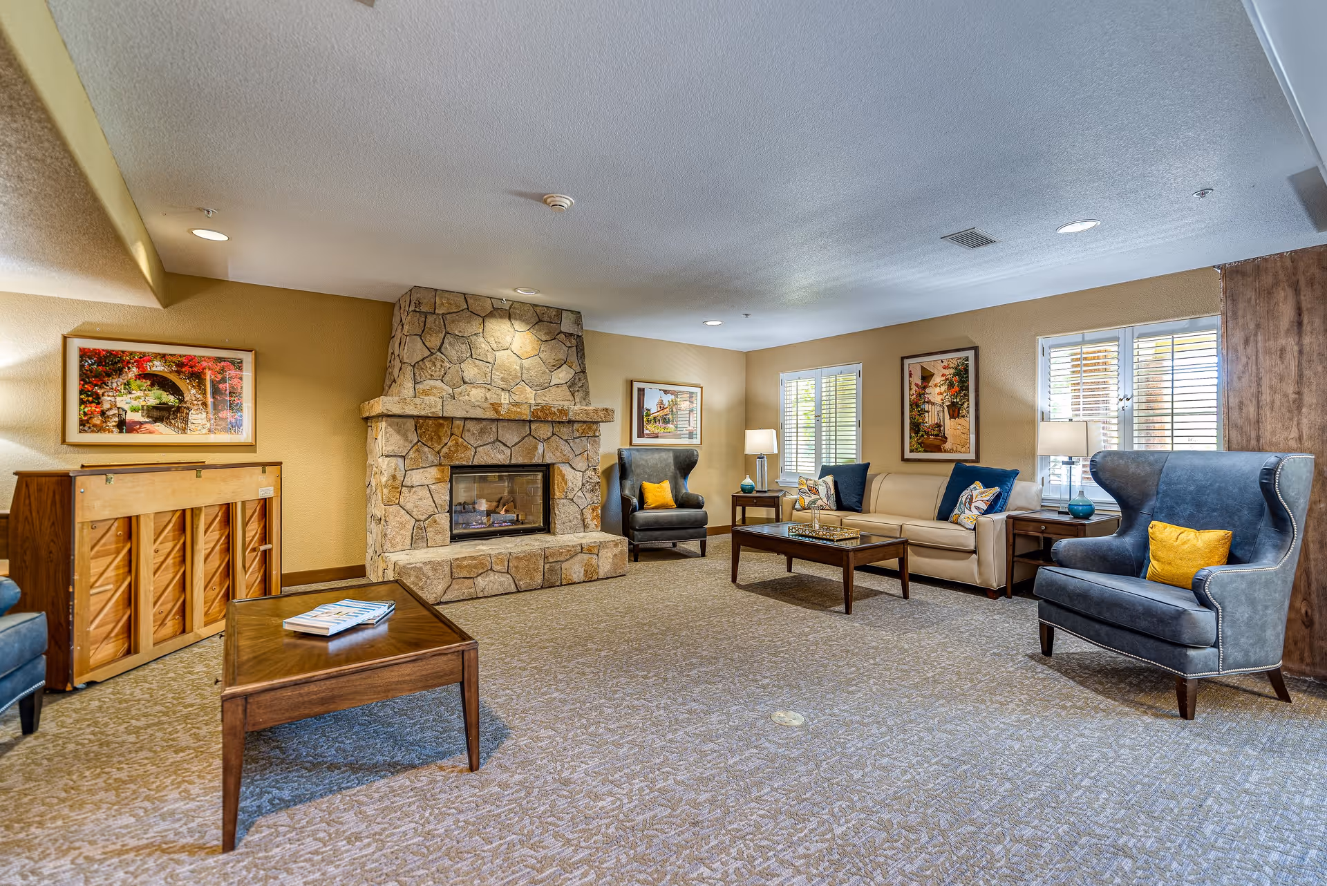 A cozy senior living room featuring a stone fireplace in the center, flanked by two blue armchairs with yellow accent pillows. A beige sofa with blue and patterned pillows sits against the wall with two windows and framed artwork. A wooden coffee table with magazines is in the foreground, and a piano is positioned against the left wall under a colorful framed picture.