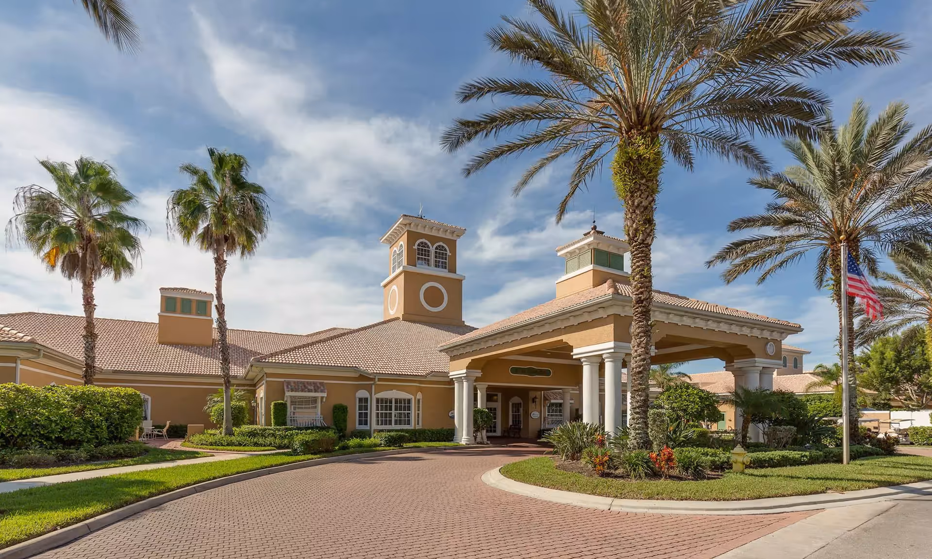 Exterior view of Aston Gardens At Pelican Marsh senior living facility with a covered entrance, palm trees, well-maintained landscaping, and an American flag on a flagpole under a partly cloudy sky.