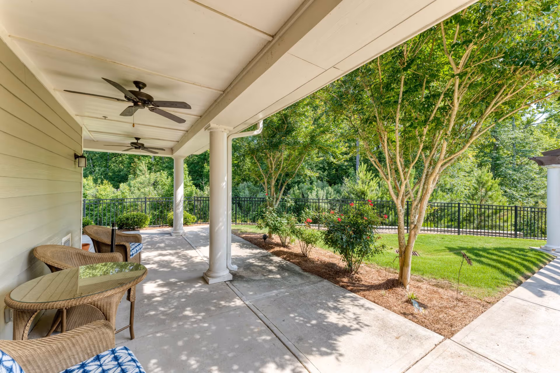 Covered outdoor patio area with ceiling fans, wicker chairs with blue patterned cushions, and a glass-top table. The patio overlooks a landscaped garden with green grass, bushes, trees, and a black metal fence.