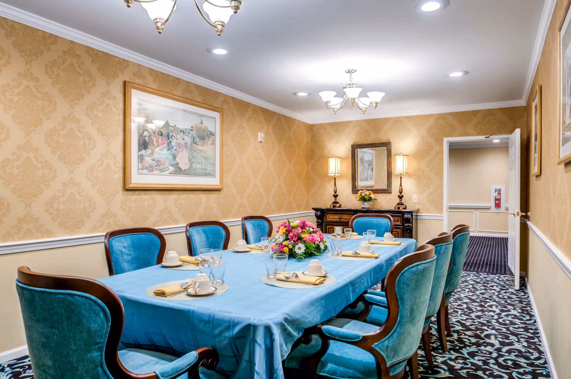 A formal dining room with a long rectangular table covered with a blue tablecloth. The table is set with white cups, saucers, glasses, and beige napkins. There is a colorful floral centerpiece in the middle of the table. The room has patterned wallpaper in beige tones, a framed painting on the wall, two table lamps on a sideboard, and a patterned carpet. The door to the room is open, showing a hallway with beige walls and a fire extinguisher.