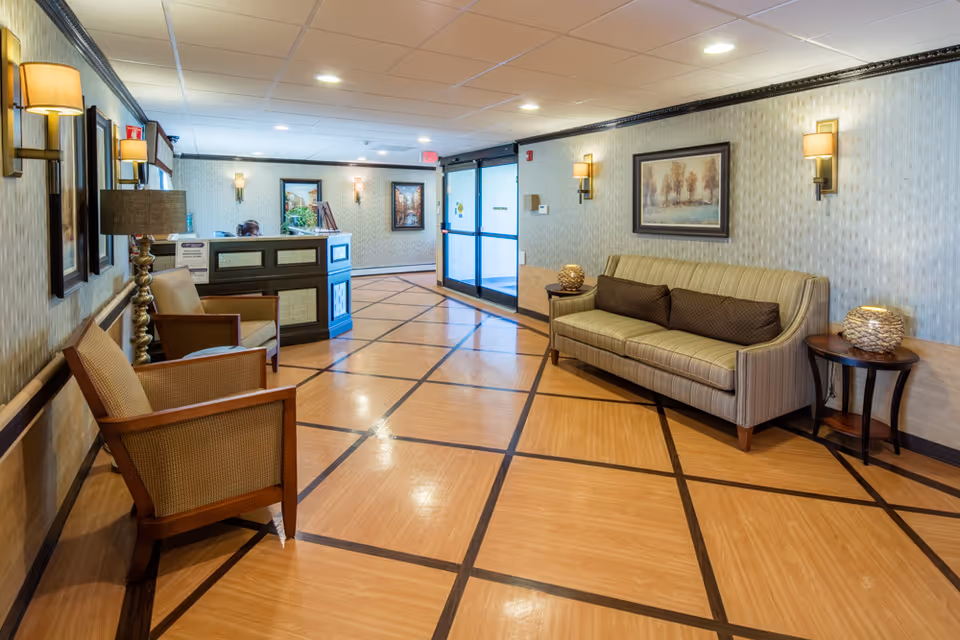 A well-lit reception area in Alpine Rehabilitation & Nursing Center featuring a wooden floor with a geometric pattern, a reception desk with a person behind it, two armchairs on the left, a striped sofa with three brown cushions on the right, side tables with decorative lamps, framed artwork on the walls, and wall-mounted light fixtures.