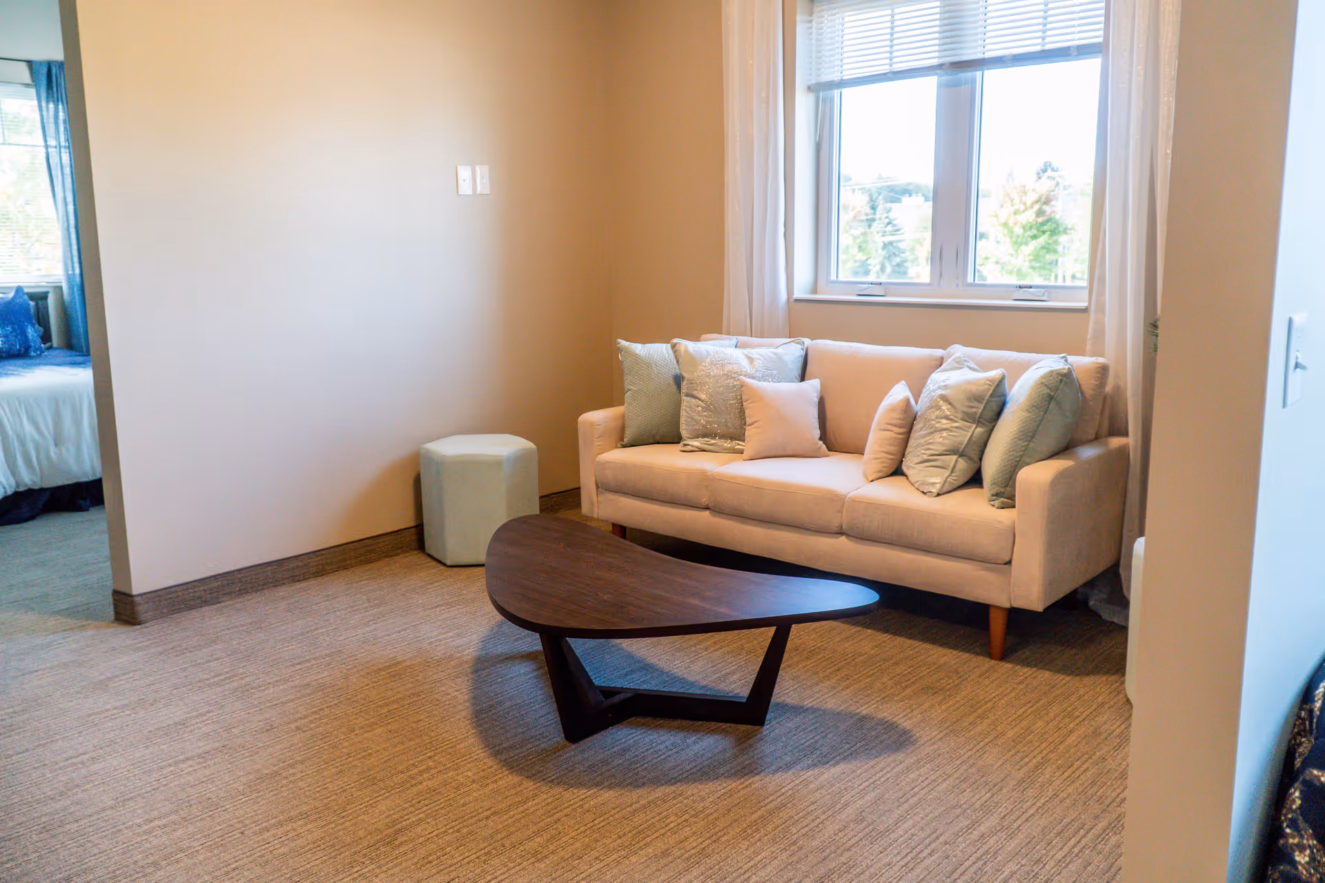 A cozy living room area with a beige sofa adorned with multiple cushions in pastel colors, a uniquely shaped dark wooden coffee table, a small light-colored ottoman, and a window with white blinds letting in natural light. A bedroom with a bed and blue pillows is partially visible through an open doorway.