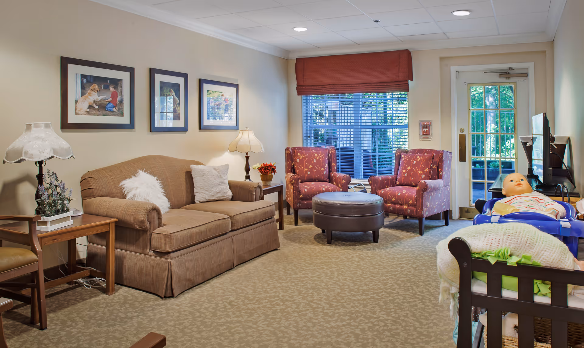 A cozy living room with a brown sofa adorned with two pillows, two red patterned armchairs, a round brown ottoman, and a side table with a lamp and flowers. There are three framed pictures on the wall above the sofa. A window with blinds and a red valance lets in natural light. A door with glass panels is visible in the background. In the foreground, there is a baby doll in a blue walker and a crib with a white knitted blanket.