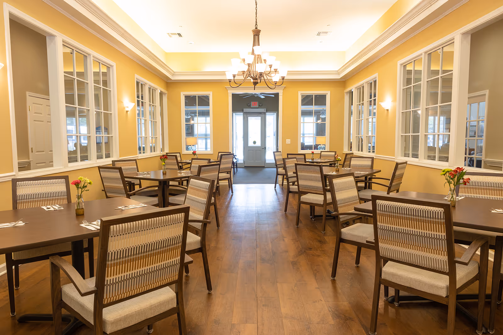 A bright and clean dining room with wooden floors, yellow walls, and multiple tables each surrounded by chairs. Each table has a small vase with flowers and place settings with forks and knives. Large windows with white trim line the walls, and a chandelier hangs from the ceiling. At the far end, there is a door with windows on either side.
