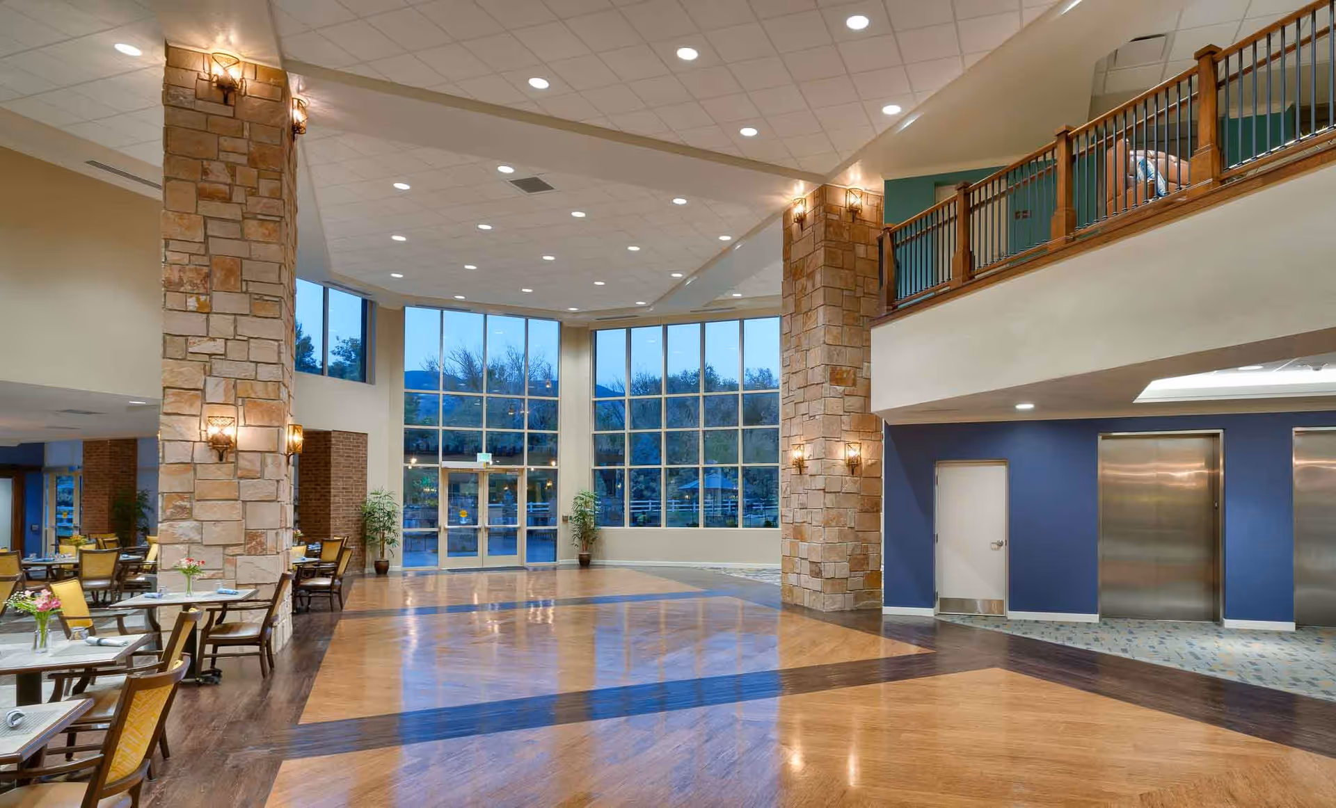 Spacious, well-lit senior living facility lobby with large floor-to-ceiling windows, stone columns, seating area, and elevators.