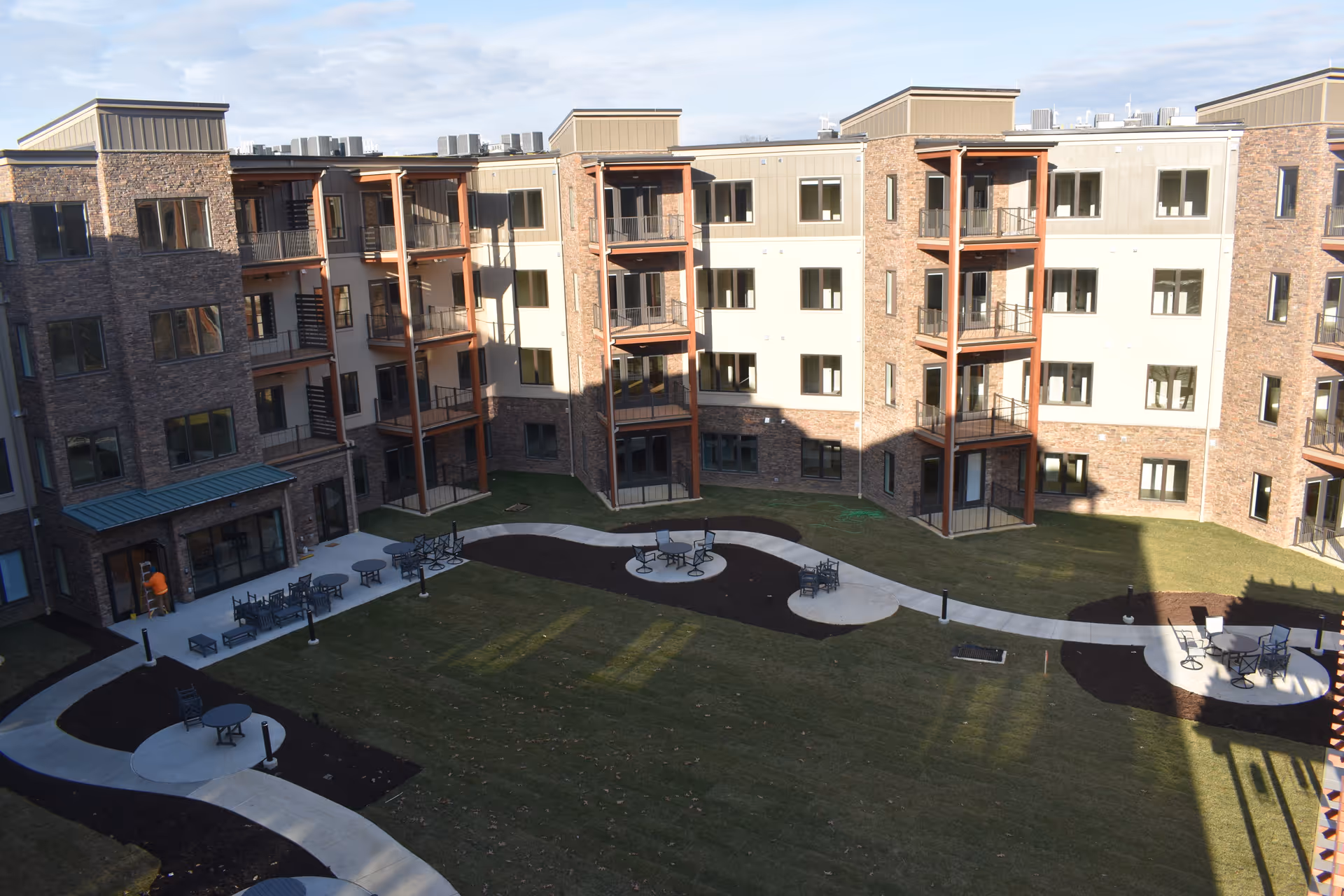 Central landscaped courtyard with winding concrete paths, patio tables and chairs, and a surrounding multi-story brick apartment building with balconies.