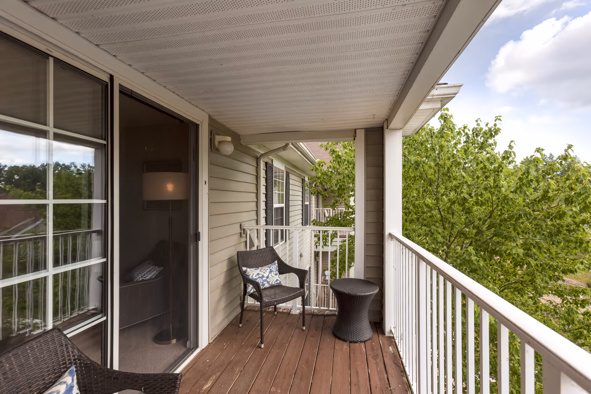 A covered balcony with wooden flooring featuring two dark wicker chairs with patterned cushions and a matching small round table. The balcony has white railings and overlooks green trees under a partly cloudy sky. A sliding glass door leads to an interior room with a floor lamp and sofa visible inside.