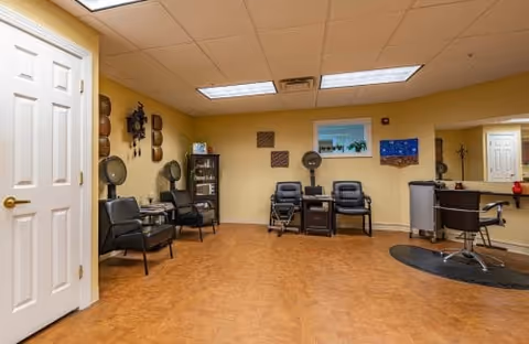 A room with yellow walls and a brown floor featuring several black chairs arranged along the walls. There is a small cabinet with decorative items, wall art including masks and framed pictures, and a counter with a chair on a black mat. The ceiling has fluorescent lights.