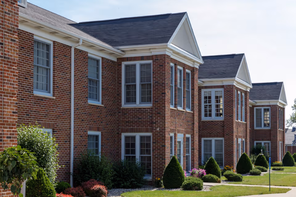Front exterior of a red brick, multi-unit retirement community building with white-trimmed windows and manicured shrubs.