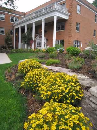 Brick senior living building with white columns, balcony, and yellow flowering garden in foreground.