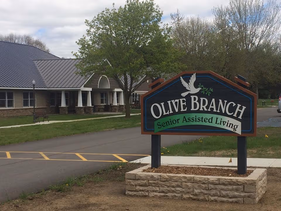 Outdoor view of the entrance area of Olive Branch Senior Assisted Living facility, featuring a large sign with a white dove holding an olive branch and the facility name. The building with a metal roof and a tree are visible in the background.