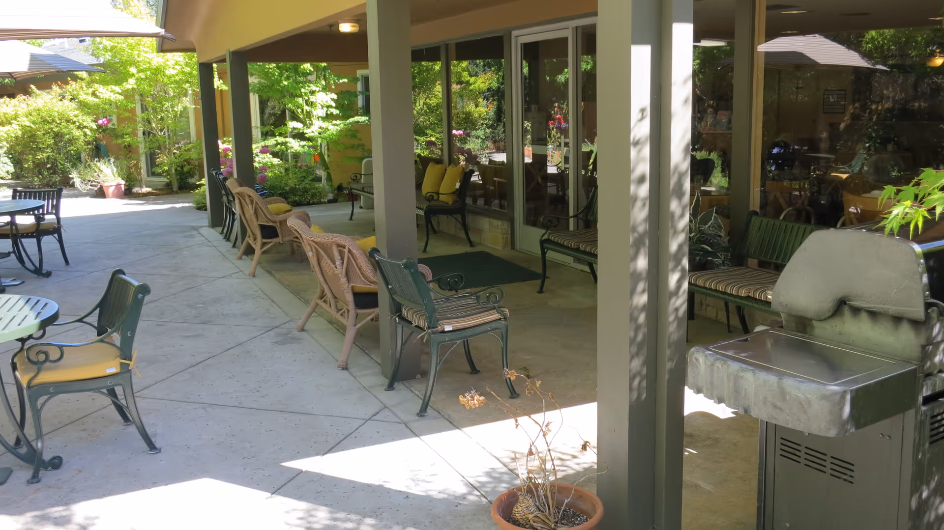 Outdoor patio area at Creekwood Care Facility with multiple chairs and benches arranged along a covered walkway. There is a barbecue grill on the right side and greenery including bushes and trees surrounding the space. Glass doors lead inside the building.