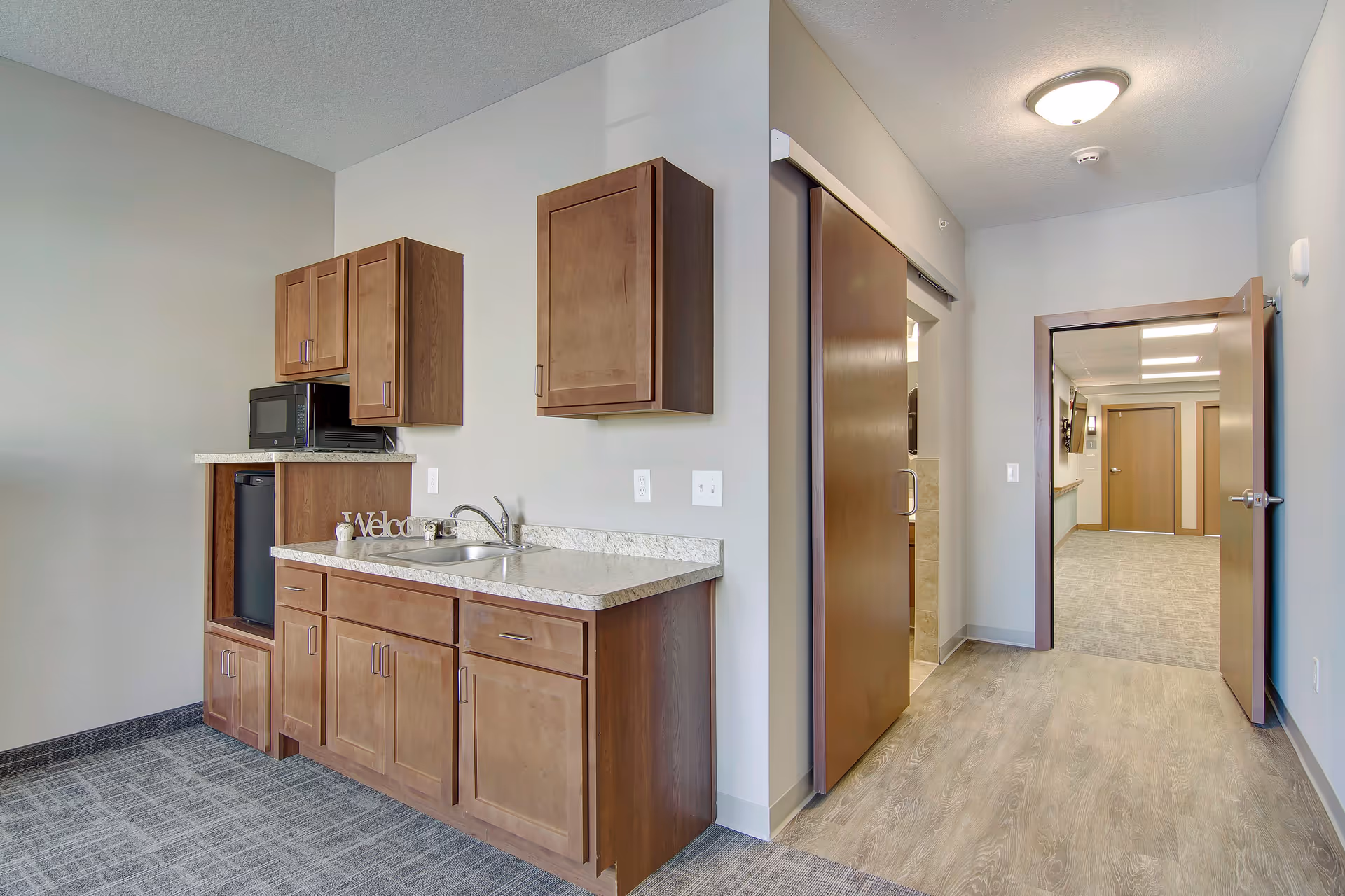 Interior view of a kitchenette area in a senior living facility with wooden cabinets, a countertop with a sink, a microwave, and a mini refrigerator. The floor transitions from carpet to wood-like flooring leading to an open doorway and a hallway with additional doors and lighting fixtures.