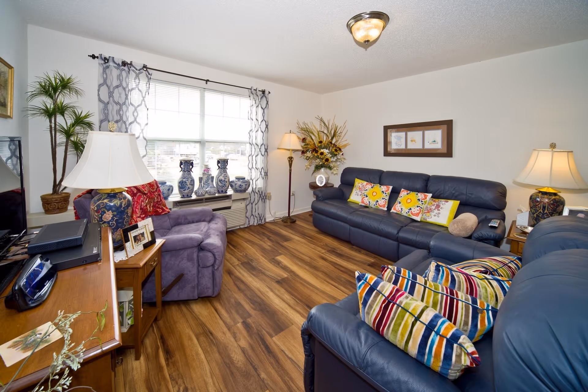 A cozy living room with wood flooring, a large window with patterned curtains, a purple armchair, two blue leather sofas adorned with colorful pillows, two table lamps with decorative bases, a floor lamp, a wooden side table with framed photos, and a collection of blue and white ceramic vases on the window sill.