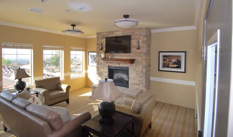 A cozy living room with beige walls and carpeted floor featuring a stone fireplace with a mounted flat-screen TV above it. The room has large windows letting in natural light, a brown leather sofa, two patterned armchairs, a dark wooden side table with a lamp, and framed artwork on the wall.
