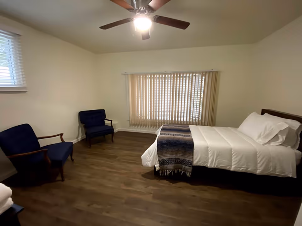 A simple bedroom with a bed made with white linens and a patterned throw blanket. Two blue upholstered chairs with wooden arms are placed against the wall. The room has wood flooring, a ceiling fan with a light, and windows with blinds and vertical curtains.