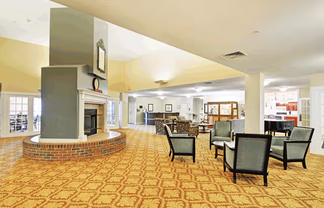 Spacious senior living facility common area with patterned carpet, a large central fireplace with a clock and mirror above it, multiple seating arrangements including armchairs and sofas, and a reception desk in the background. The room is well-lit with natural and artificial light.