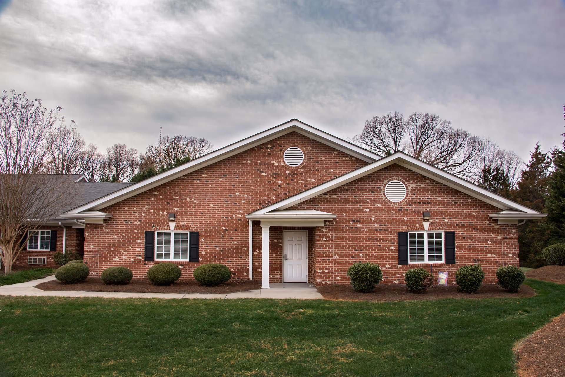 Front exterior view of a single-story brick building with a white door centered under a small covered porch. The building has two windows with black shutters on either side of the door, neatly trimmed bushes in front, and a well-maintained lawn. Trees without leaves are visible in the background under a cloudy sky.