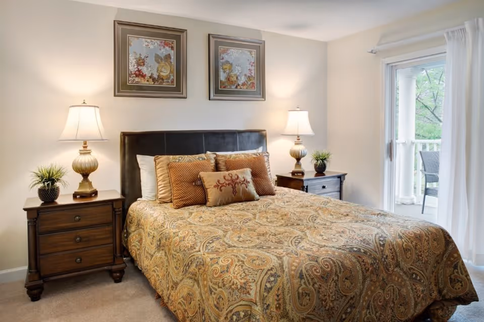 A cozy bedroom featuring a large bed with a patterned bedspread and multiple decorative pillows. On each side of the bed are wooden nightstands with matching lamps and small potted plants. Above the bed, two framed floral artworks hang on the wall. To the right, a sliding glass door with white curtains opens to a balcony with outdoor seating and greenery visible outside.