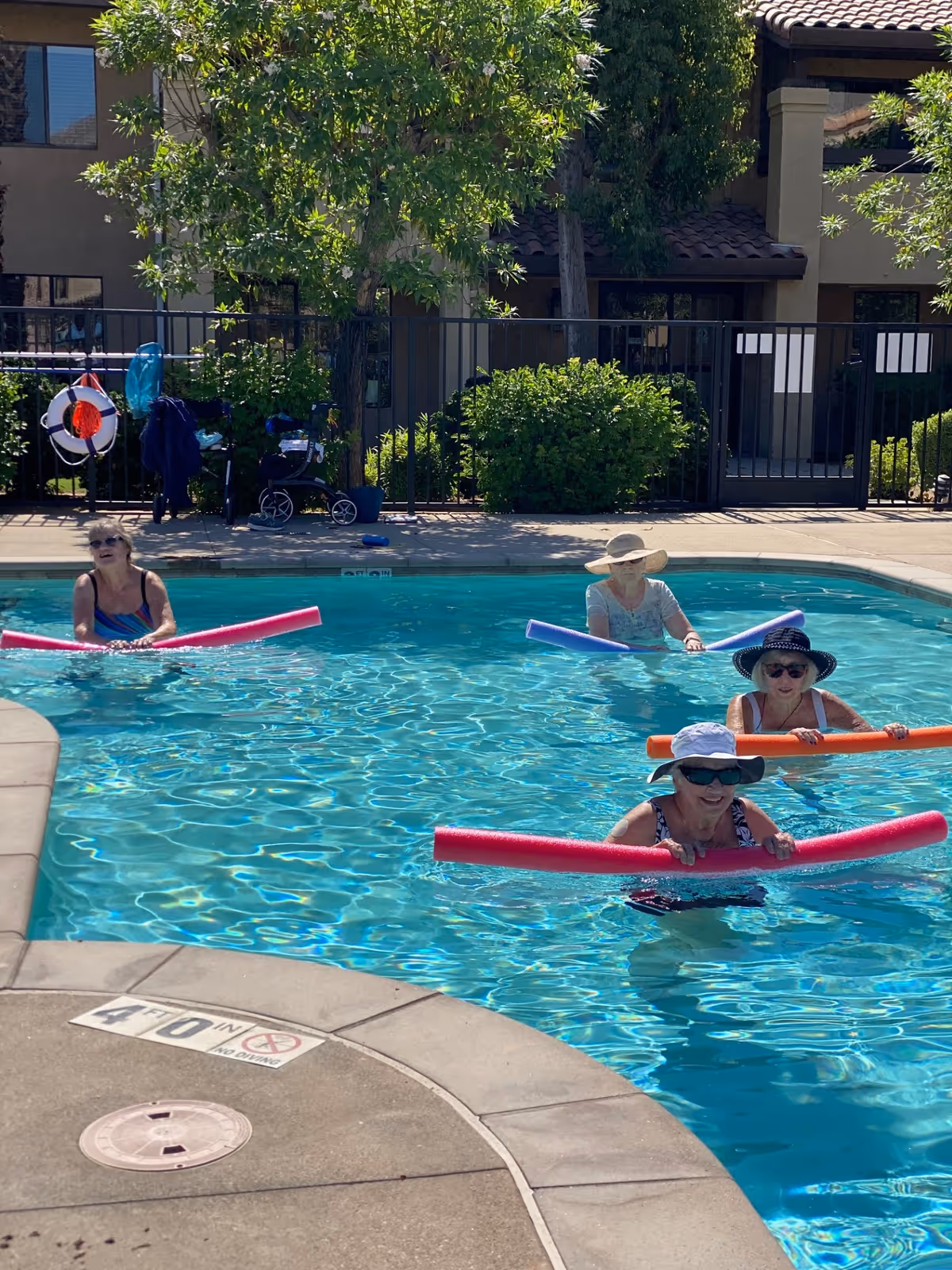 Four elderly women in a swimming pool holding colorful pool noodles, with a building and greenery in the background. The women are wearing sun hats and sunglasses, enjoying a sunny day in the water.