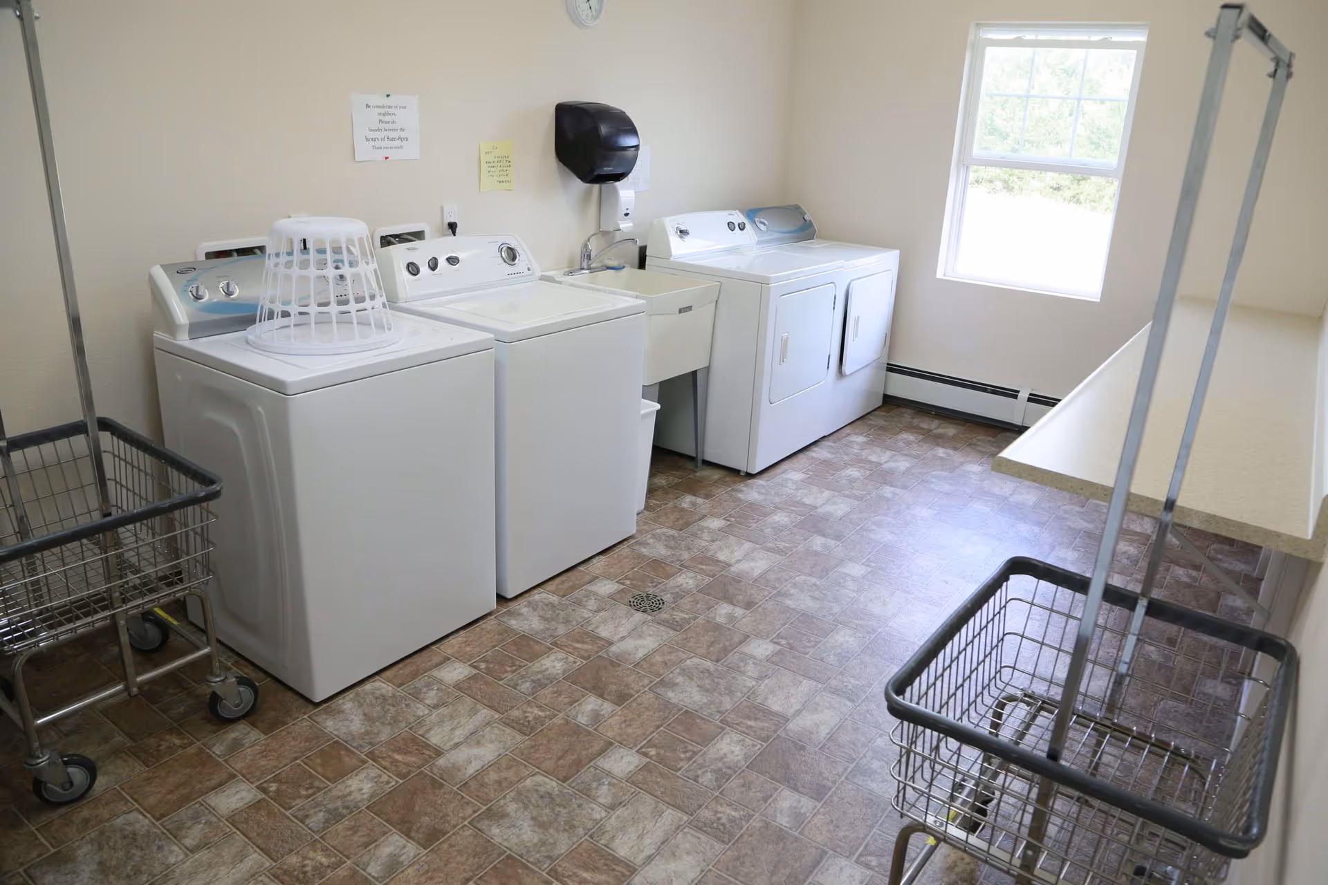 A laundry room with two washing machines and two dryers lined up against a wall. There is a utility sink between the washers and dryers. Two metal laundry carts with wheels are visible, one in the foreground and one to the left. The room has a window letting in natural light and a tiled floor.