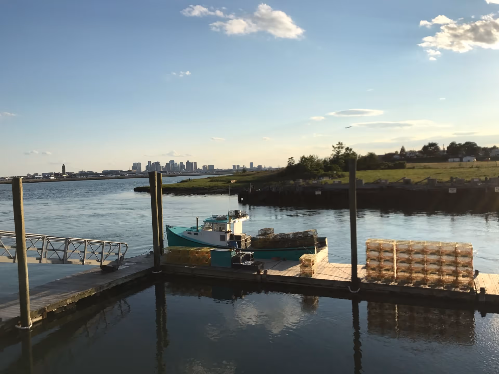 A calm waterfront scene with a small dock and a boat loaded with crates. The water reflects the sky and dock posts. In the background, there is a grassy area and a distant city skyline under a clear blue sky with a few clouds.
