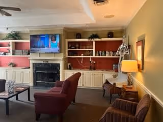 A cozy living room with beige walls and built-in white shelves with red backing. The shelves hold books and decorative items. A flat-screen TV is mounted above a black fireplace. There are two upholstered chairs and a sofa arranged around a coffee table. A floor lamp and a table lamp provide warm lighting.