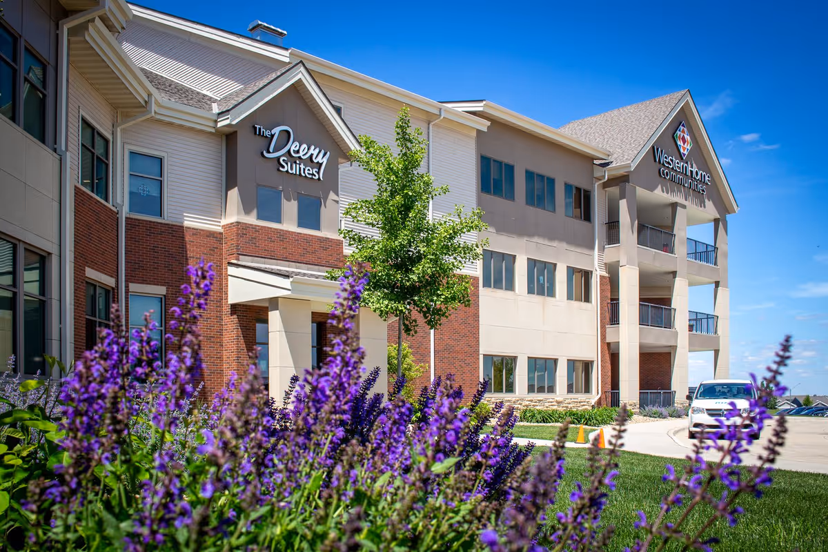 Front exterior of the Western Home Communities building with purple flowers in the foreground and signage for The Deery Suites.
