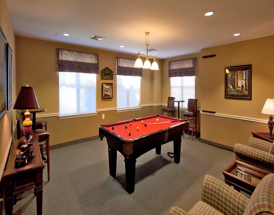 A cozy game room with a red felt pool table in the center. The room has beige walls, three windows with patterned valances, and recessed ceiling lights. There are two high chairs and a small table near the windows, a plaid armchair and side table with a lamp, a wooden console table with a lamp and decorative items, and framed artwork on the walls.