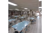 Large institutional dining room with rows of pale blue-topped tables and white metal chairs on a tiled floor under a drop ceiling with fluorescent lights.