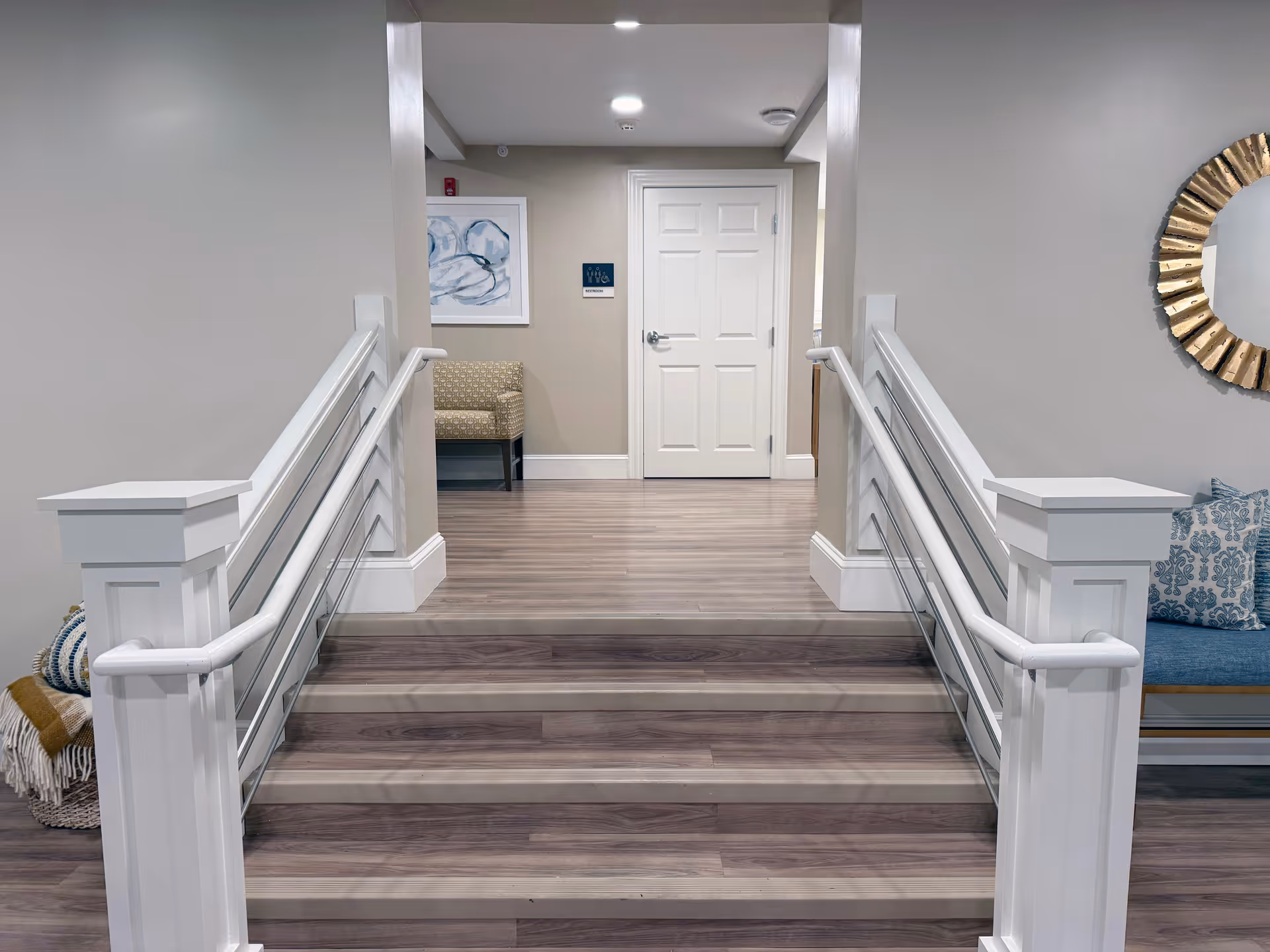 Interior view of a senior living facility hallway with wooden stairs leading up to a white door. The walls are painted light gray, with white railings on both sides of the stairs. There is a beige patterned chair and framed artwork on the left side, and a blue cushioned bench with patterned pillows and a round decorative mirror on the right side.