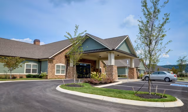 Exterior view of Emory Ridge senior living facility showing the main entrance with a covered drop-off area, stone and green siding facade, landscaped greenery, and a parking lot with a silver car parked near the entrance.