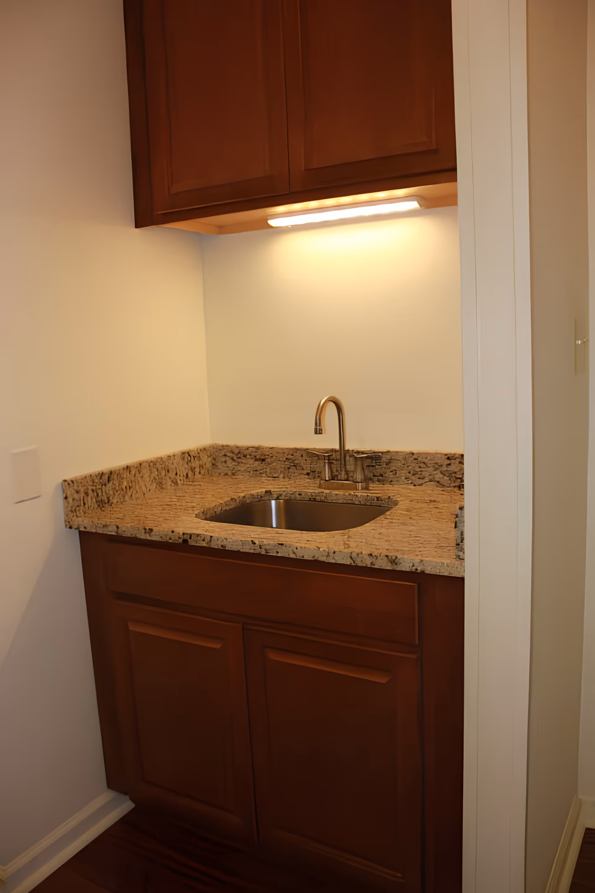 Small kitchenette with a granite countertop, stainless steel sink and faucet, and wooden cabinets above and below.