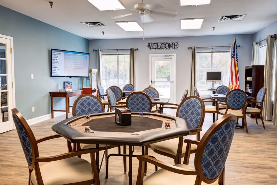A bright and spacious common area with multiple round tables and blue cushioned chairs arranged around them. One table in the foreground is a poker table with poker chips on it. The room has large windows with beige curtains, a wall-mounted TV, a welcome sign above a door, and an American flag in the corner.