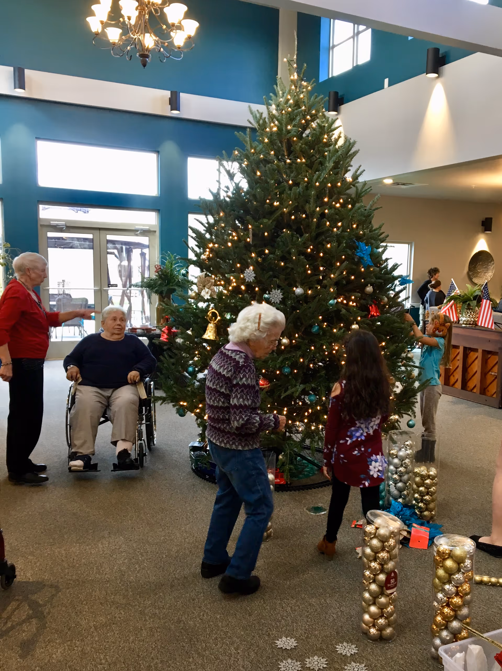 Residents decorate a large Christmas tree in a spacious assisted living facility lobby.