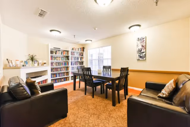 A cozy common area in Sabine Place featuring two black leather sofas with decorative pillows facing each other, a dark wooden table with four chairs in the center, a built-in bookshelf filled with books, a window letting in natural light, and a wall-mounted floral artwork.
