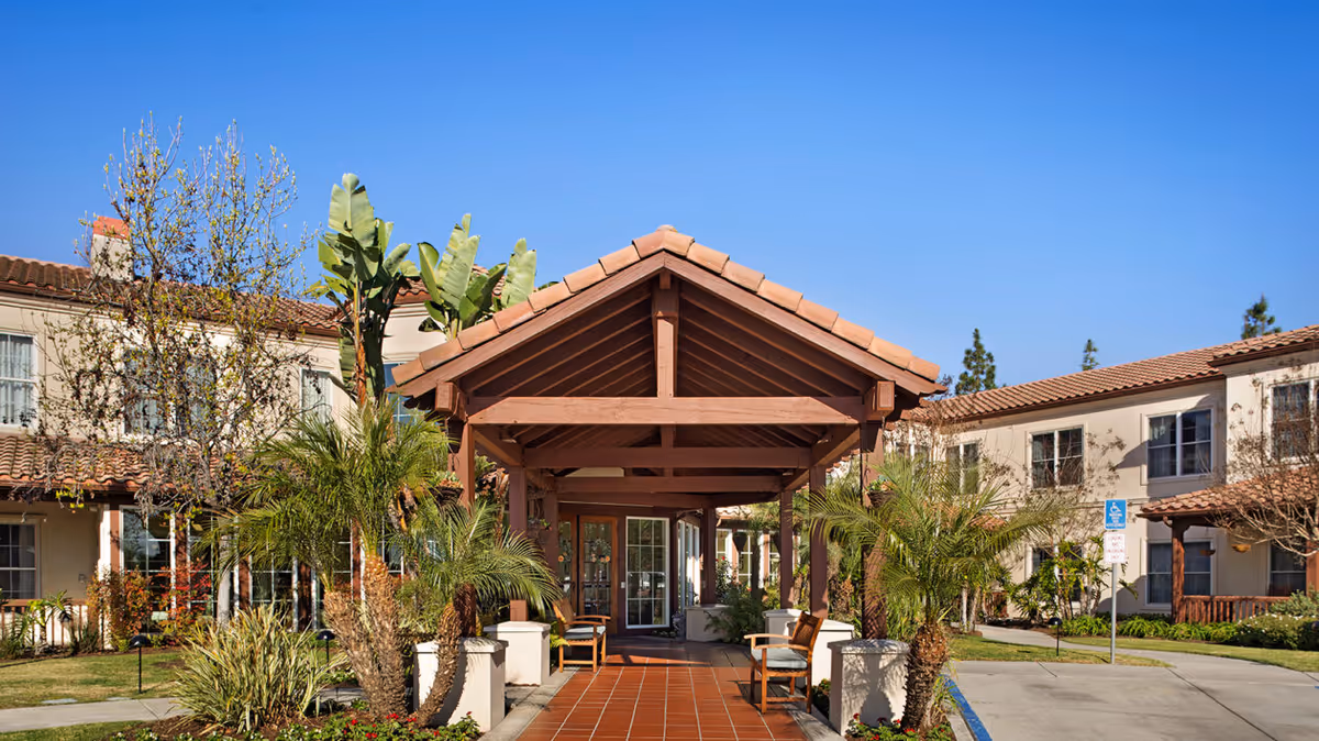 Outdoor covered walkway entrance to a senior living facility with tiled roof and wooden beams, surrounded by palm trees and other greenery, with two-story beige buildings on either side under a clear blue sky.